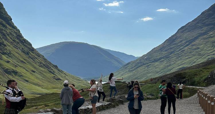 Groups of visitors pause for photos in a dramatic Scottish valley of green peaks.