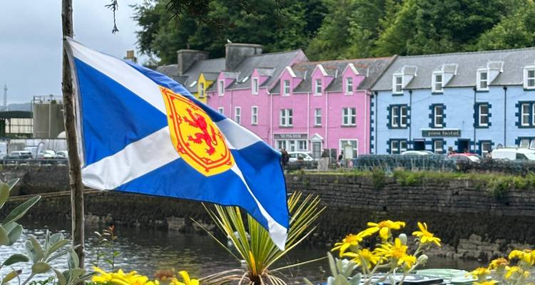 Un drapeau écossais se détache sur des cottages riverains peints en couleurs pastel et un environnement verdoyant.