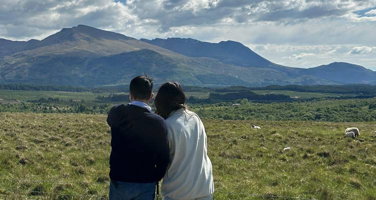 Un jeune couple se tient côte à côte, admirant les pics montagneux lointains et les moutons qui paissent sur une plaine ensoleillée.