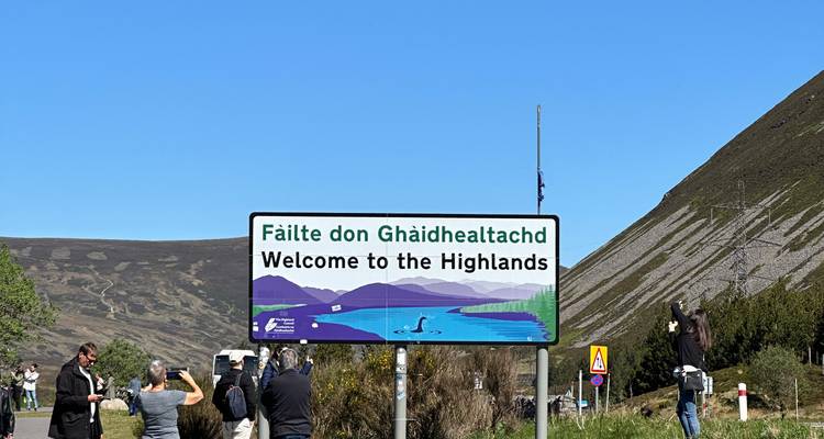 Les voyageurs prennent des photos à côté d'un panneau routier coloré « Bienvenue aux Highlands » sous un soleil éclatant.