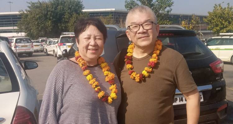 Lachend stel met welkomstslinger van goudsbloemen dat in een zonnige parkeerplaats naast auto's staat.