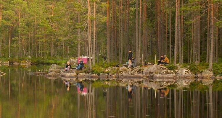 People sitting by a calm forest lake surrounded by tall trees.