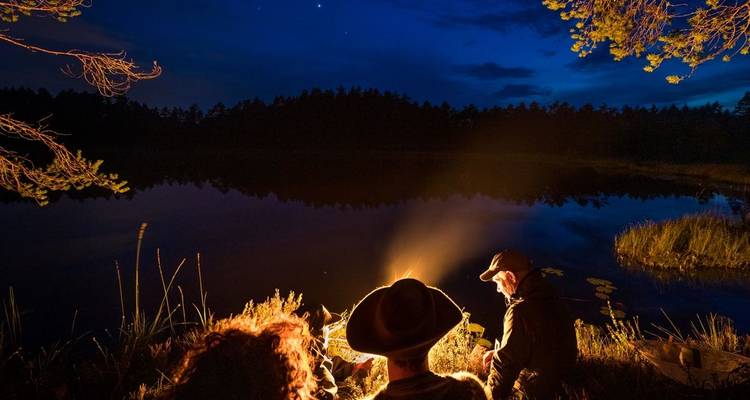 Un groupe de personnes autour d'un feu de camp la nuit, avec un lac et une forêt en arrière-plan.