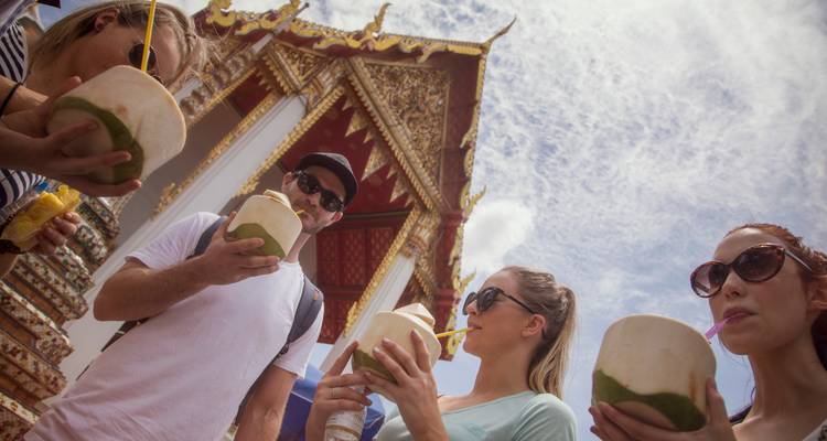 Los viajeros jóvenes beben cocos frescos afuera de un templo tailandés ornado bajo un cielo brillante.