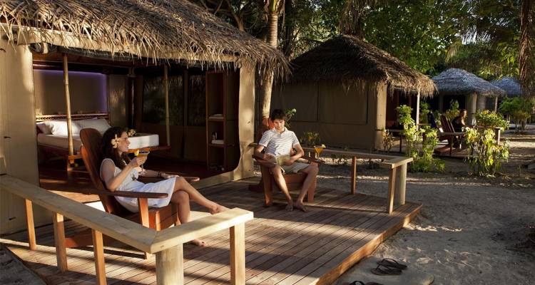 Un couple déguste des boissons sur la terrasse en bois d'un bungalow en bord de plage au toit de chaume entouré de verdure.