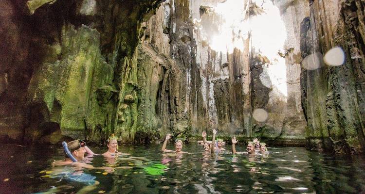 Les voyageurs agitent la main en nageant dans un bassin spectaculaire situé dans une grotte calcaire, illuminé par des rayons de lumière naturelle provenant d'en haut.