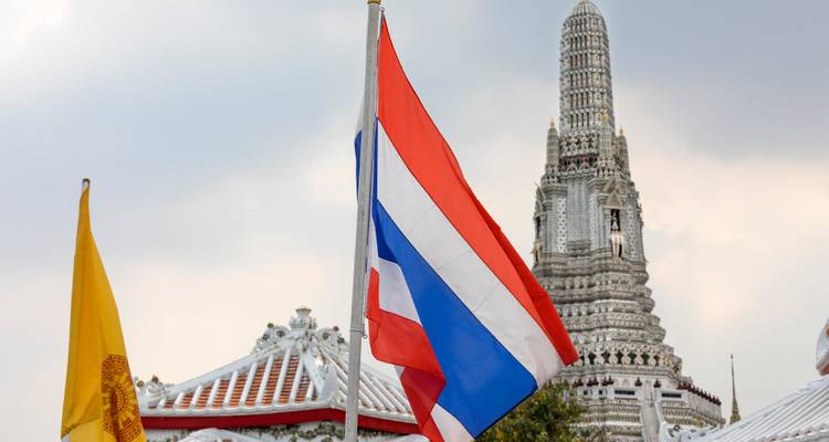 Le drapeau national thaïlandais flottant avec le prang orné du Wat Arun se dressant en arrière-plan contre un ciel nuageux.