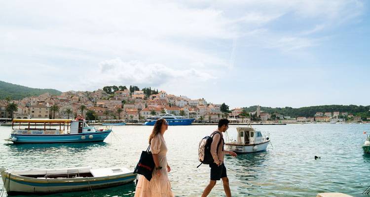 Un couple se promène le long d'un port adriatique ensoleillé avec une ville croate perchée sur une colline et des bateaux derrière eux.