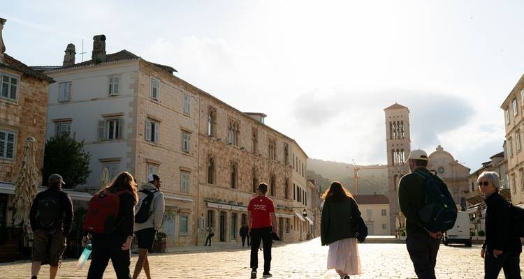 Les voyageurs explorent une place pavée de pierre baignée de soleil, bordée par des bâtiments historiques et un clocher d'église.