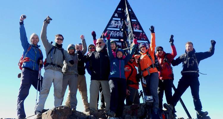 Ein triumphales Trekkingteam posiert neben der eisernen Gipfelpyramide des Mount Toubkal unter einem strahlend blauen Himmel.