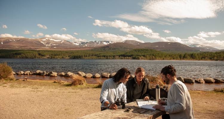 Three people sitting at a picnic table by a lake.