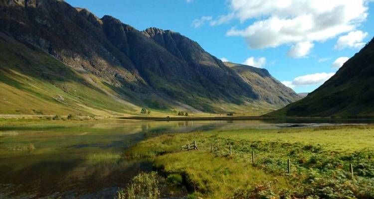 A tranquil valley with steep green mountains reflects softly in a shallow stream under scattered clouds.