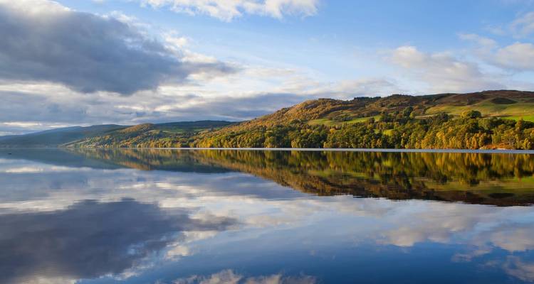 Calm lake water perfectly mirrors rolling green hills and a sky filled with mixed clouds.