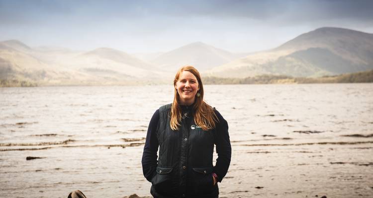 Woman standing by a loch with hills in the distance.