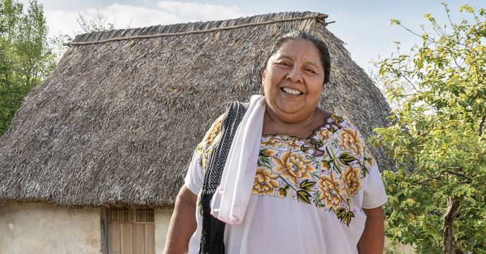 A Mexican woman wearing traditional clothes standing next to her house as if welcoming guests