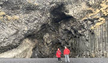 Two people in red jackets standing in front of basalt columns at a beach.