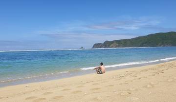 A person sitting on a sandy beach with clear blue waters and distant hills.