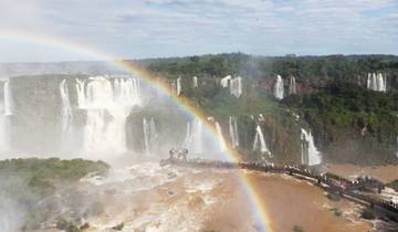 A wide view of Iguazu Falls with a visible rainbow and tourists on a walkway.