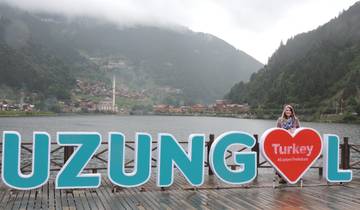 A person posing next to a large Uzungöl sign with a village and nature backdrop.