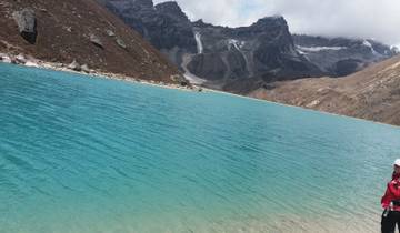 Person standing by a vibrant blue lake in a mountainous region.