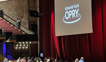 Interior of a theater with a stage and audience. 'Grand Ole Opry' displayed on a screen.