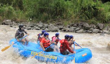 Team navigating a raft through rapids, lush riverbank visible.
