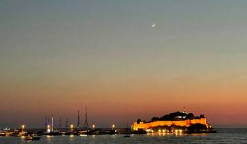 Seaside fort lit up at sunset with crescent moon.