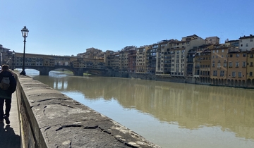 A person walking along a riverbank with historic buildings.