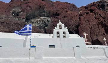 A Greek church with a prominent flag and bell tower set against a rocky landscape.