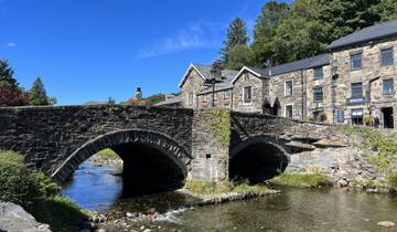 Stone bridge with arches over a gently flowing river, surrounded by historic buildings.