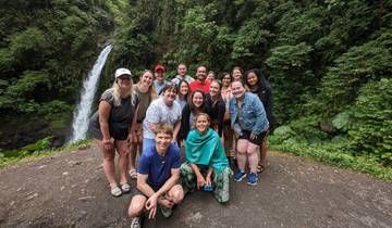 A group of tourists posing in front of a waterfall in a forest setting.
