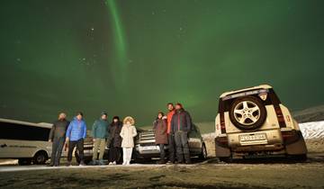 People standing under the northern lights with cars nearby.