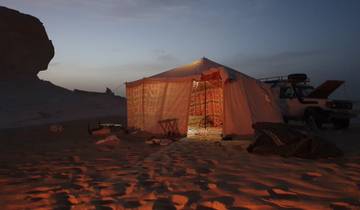 A tent lit up at night in a desert area with a vehicle nearby.