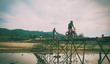Motorbikes crossing a rustic wooden bridge over a reflective water body.