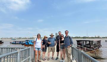 Group of people standing on a pier with boats on the river and buildings in the background.