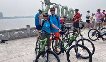 Group of cyclists posing in front of a scenic lake with a dragon sculpture.