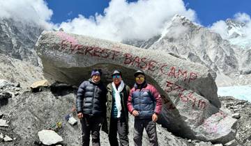 Three people standing in front of a large rock with Everest Base Camp inscription, snowy mountains in the background.