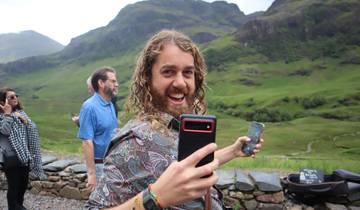 A smiling man holding two phones with a scenic green landscape in the background.