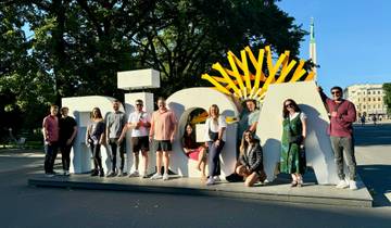 Group posing with 'Riga' lettering and cityscape background.