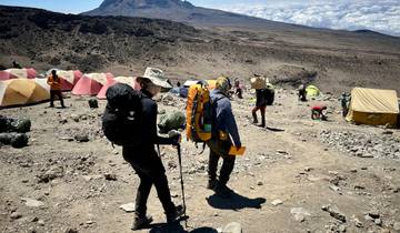 Hikers with backpacks walking past tents on a rocky terrain.