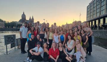 Large group posing by water during sunset with cityscape.