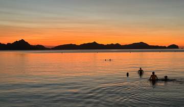 People swimming at sunset with an orange and purple sky over a mountain horizon.