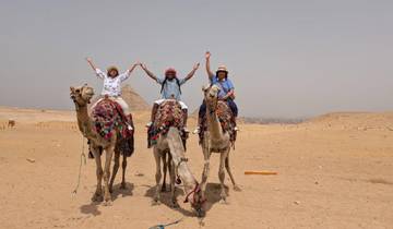 Three people riding camels in the desert with pyramids in the background.