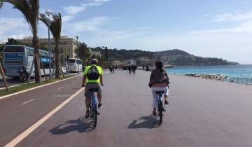 Bicyclists on a promenade by the sea with hills in the background.