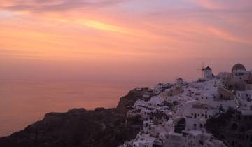 White buildings with blue domes on a cliffside overlooking a pink sunset.
