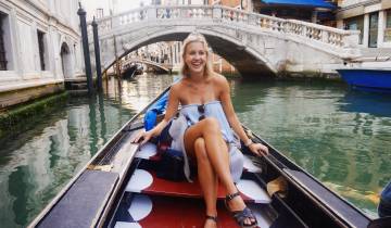 Woman sitting on a gondola in a canal with a bridge behind her.
