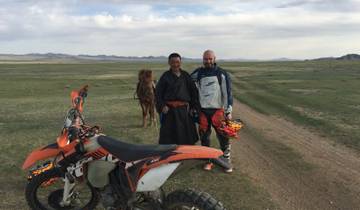 Two people standing beside motorcycles on a vast plain with a horse in the background.