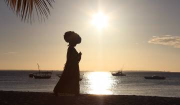 Silhouette of a person walking on the beach with boats on the water.