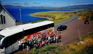 Tour group posing energetically in front of a white tour bus on a scenic coastal road.