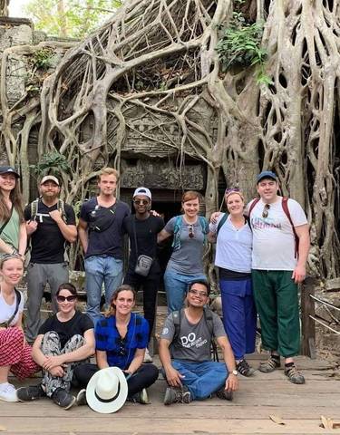 Group of tourists in front of tree roots covering temple ruins.